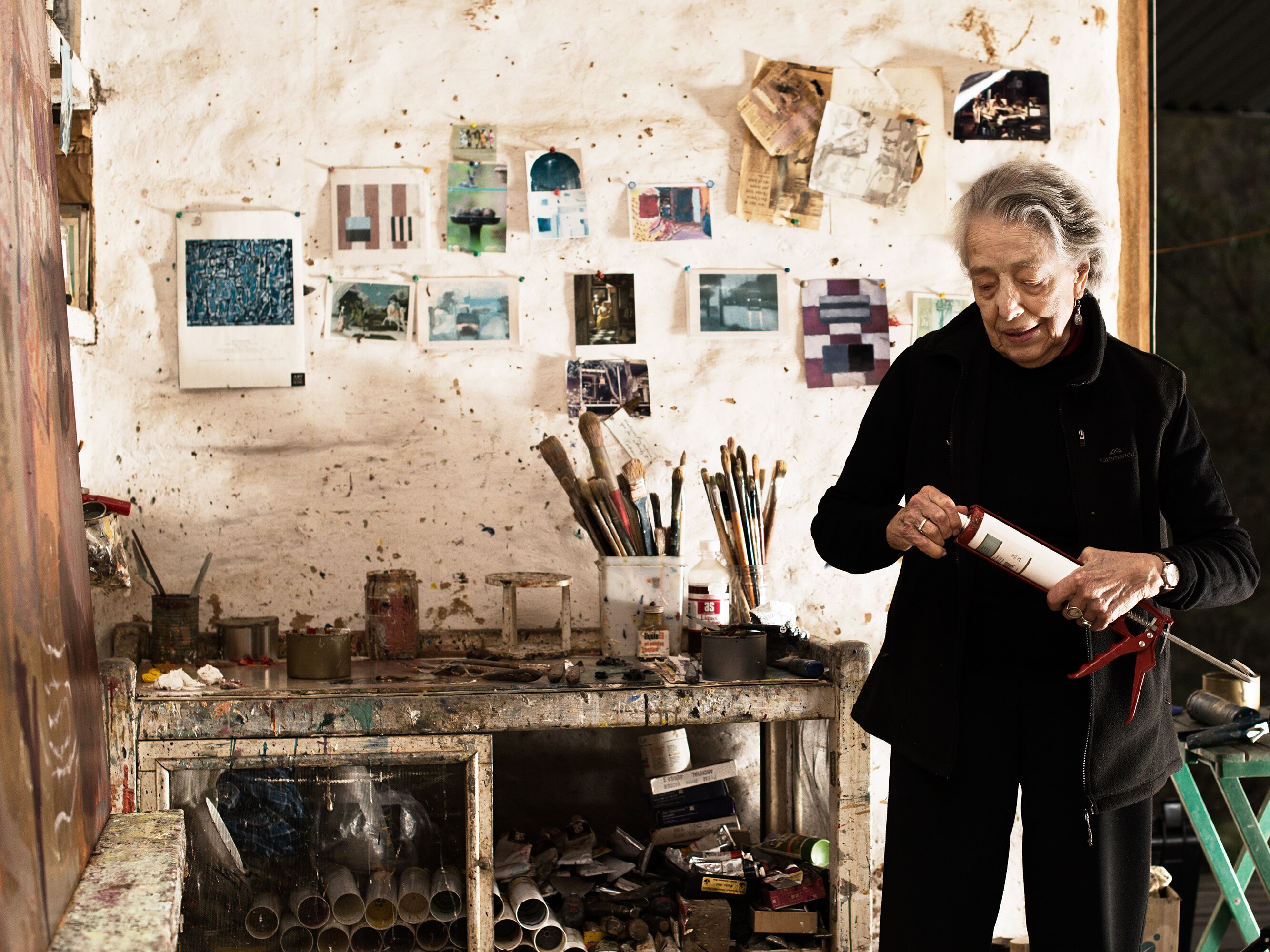 A woman stands in front of art on a wall and paintbrushes on a desk.