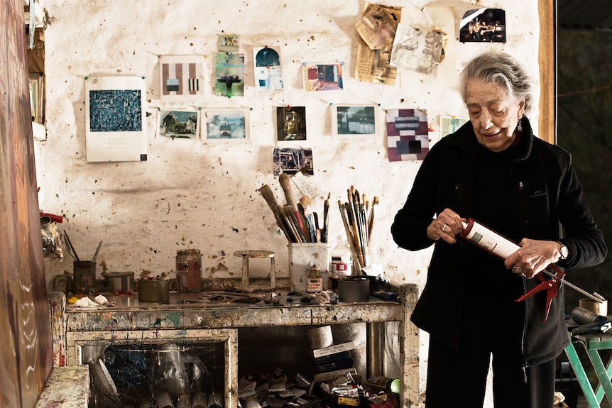 A woman stands in front of art on a wall and paintbrushes on a desk.