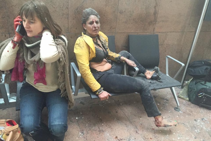 Two women sit on chairs bloodied, one woman is on the phone