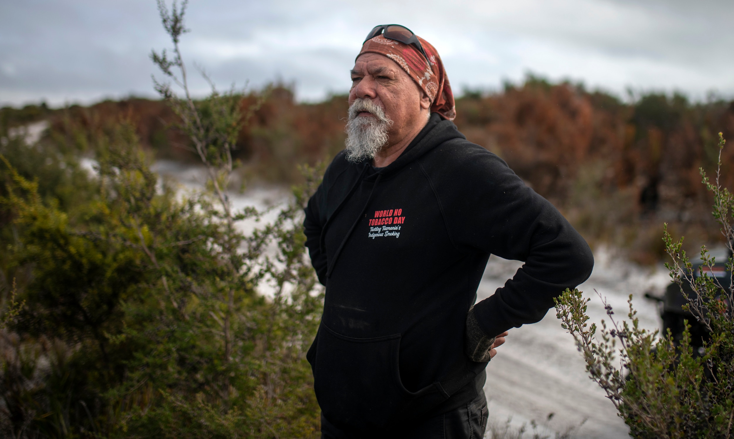 A man in a red bandana and black shirt with a grey beard stands with his hands on his hips by a sandy track in bushland. 