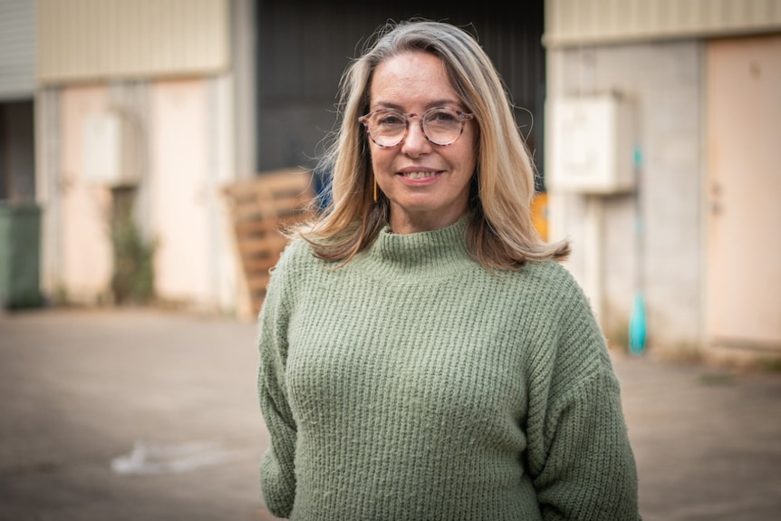 A smiling blonde woman wearing a green turtleneck jumper and glasses.