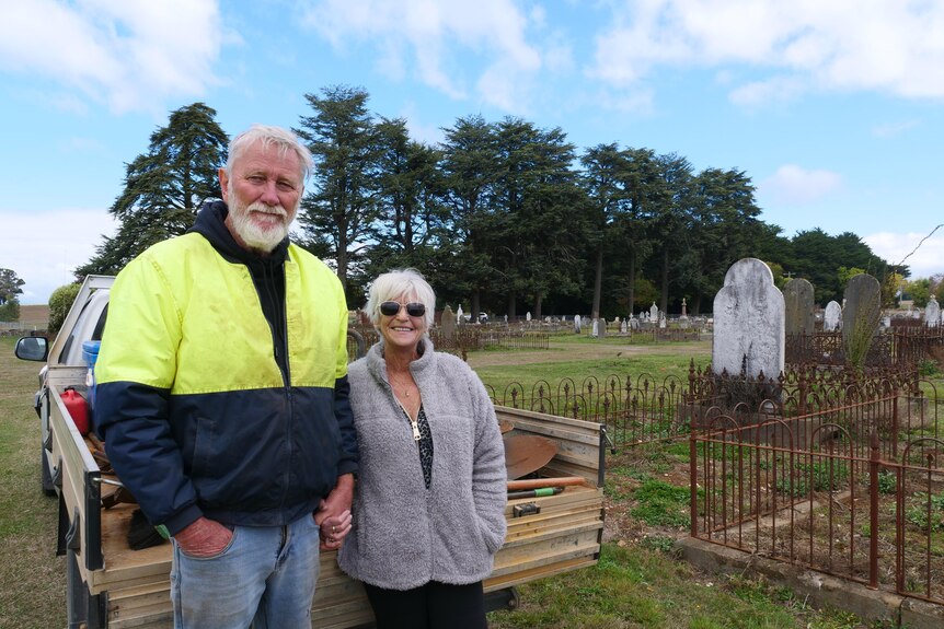 man and woman in their sixties standing and smiling in a graveyard. 