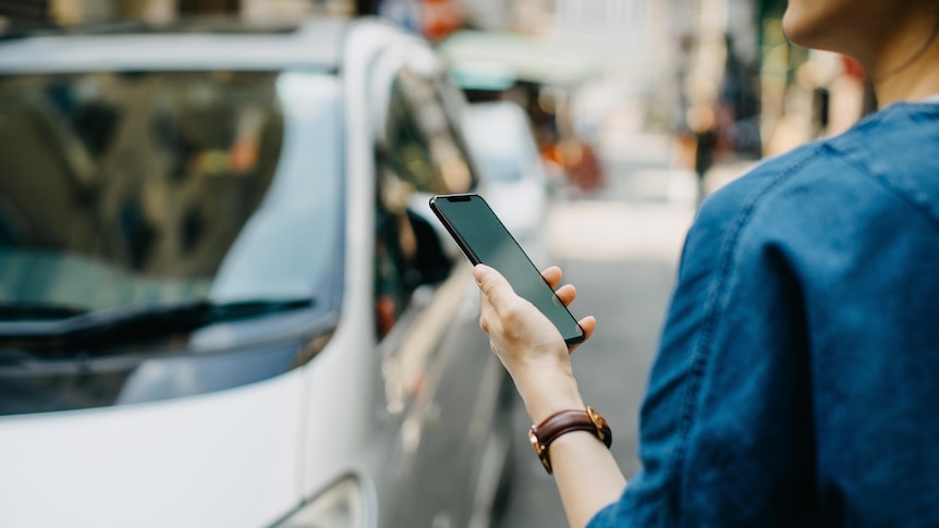 A woman, shot from behind, holding a smart phone on a street where cars are seen parked on the road.