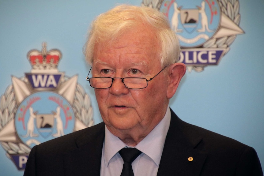 A bespectacled older man wearing and a black suit speaks in front of a police backdrop