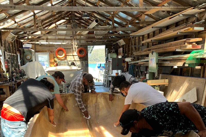 Men sanding a fibreglass boat in a small wooden boatshed