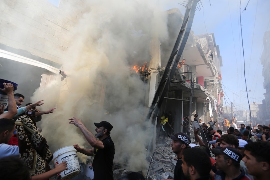 heavily damaged building surrounded by smoke while people huddle around