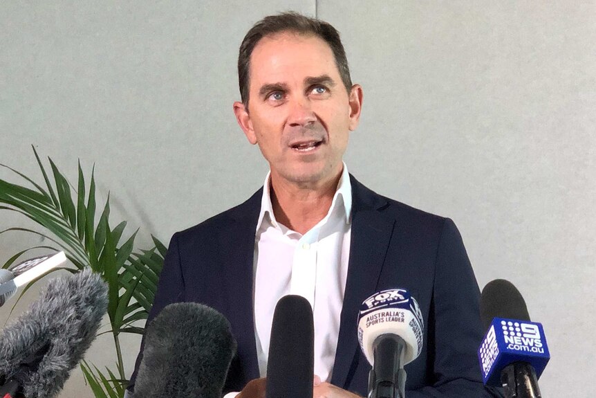 A mid shot of Justin Langer speaking indoors at a media conference in front of microphones, wearing a suit and shirt.