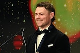 Brisbane Lions player Lachie Neale smiles while standing behind a podium with the Brownlow Medal on a screen behind him.