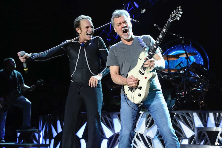 A man laughs while he holds a microphone while another man plays guitar.