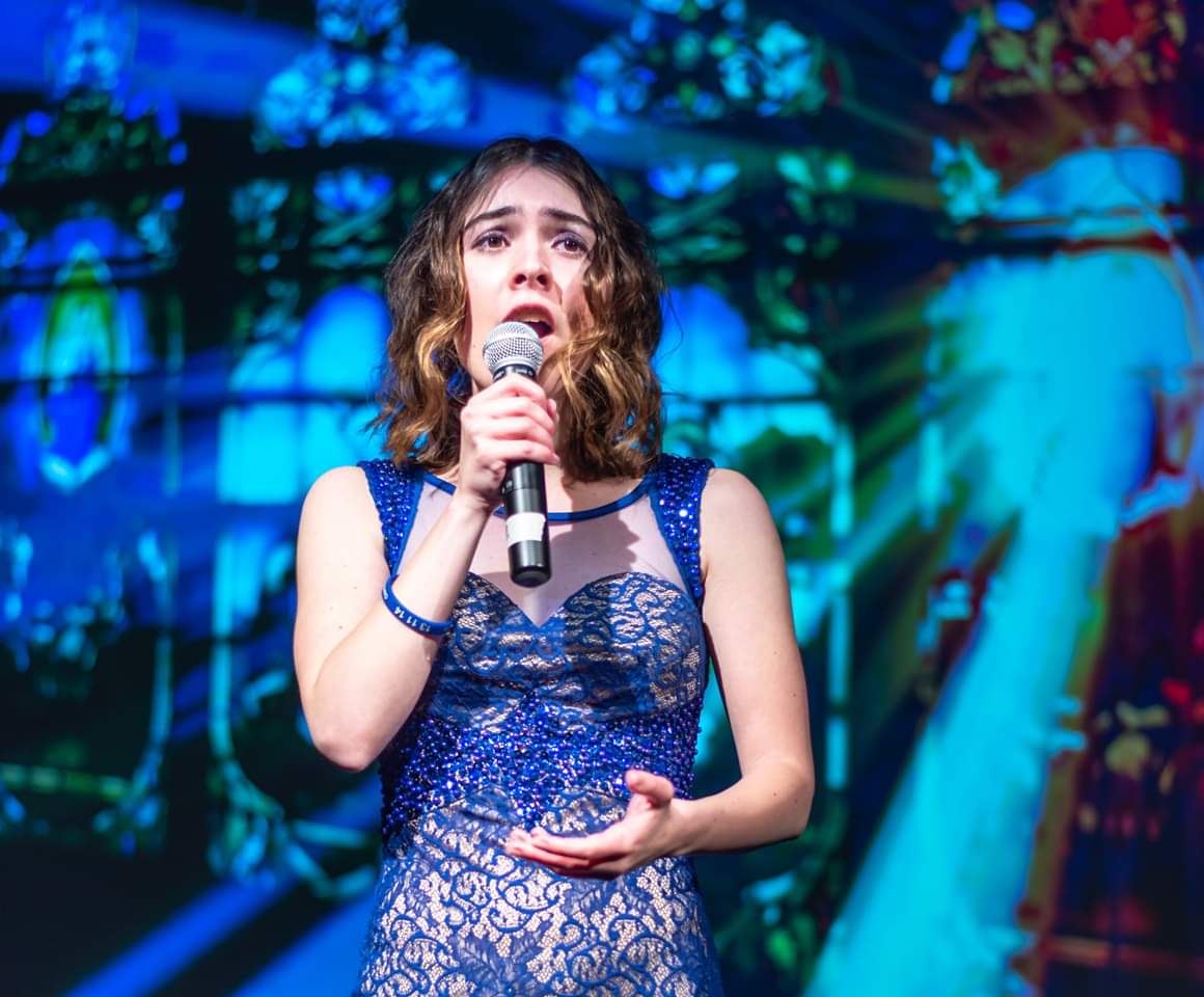 A young woman in a blue dress holds a microphone to their face as they sing behind a blue backdrop