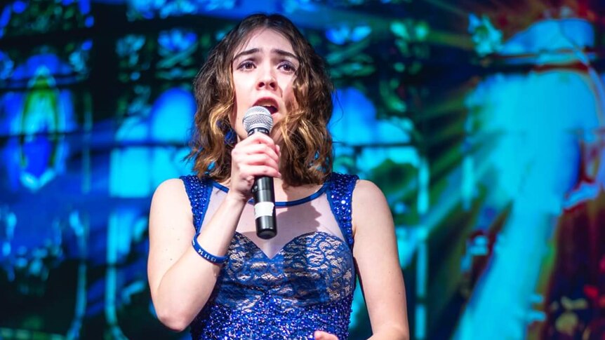 A young woman in a blue dress holds a microphone to their face as they sing behind a blue backdrop