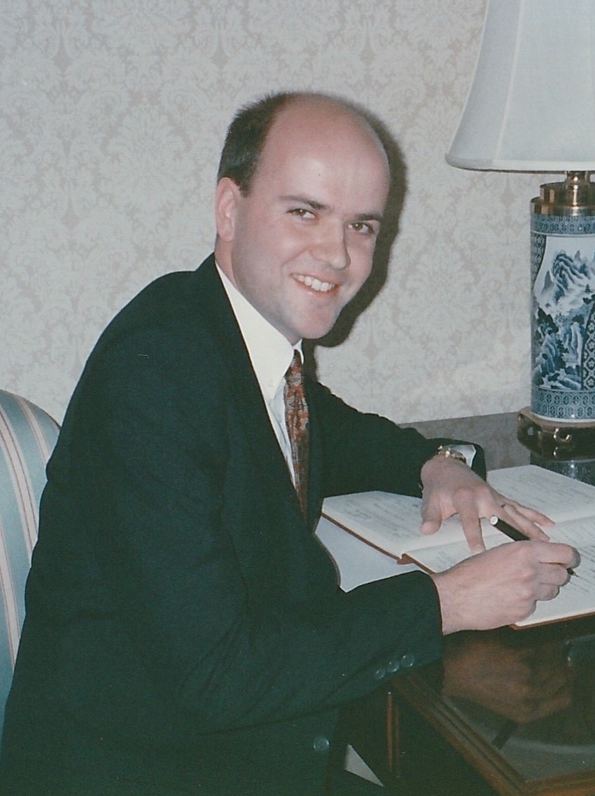 A young man wearing a suit sits at a desk and writes in a notebook.