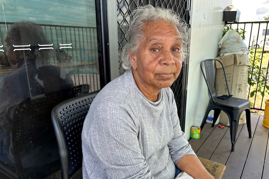 A First Nations woman with grey hair sits on a chair on a verandah, with a sad expression on her face.