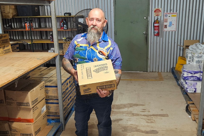 a bald man with a beard holding a carton of food in a near-empty store room