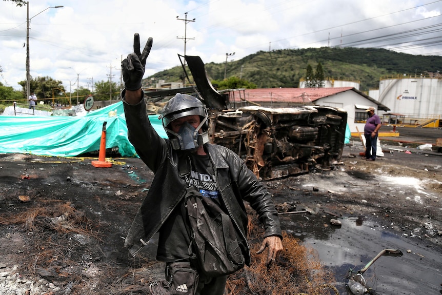 sA man flashes a victory sign near the charred remains of a car that was burned by protesters