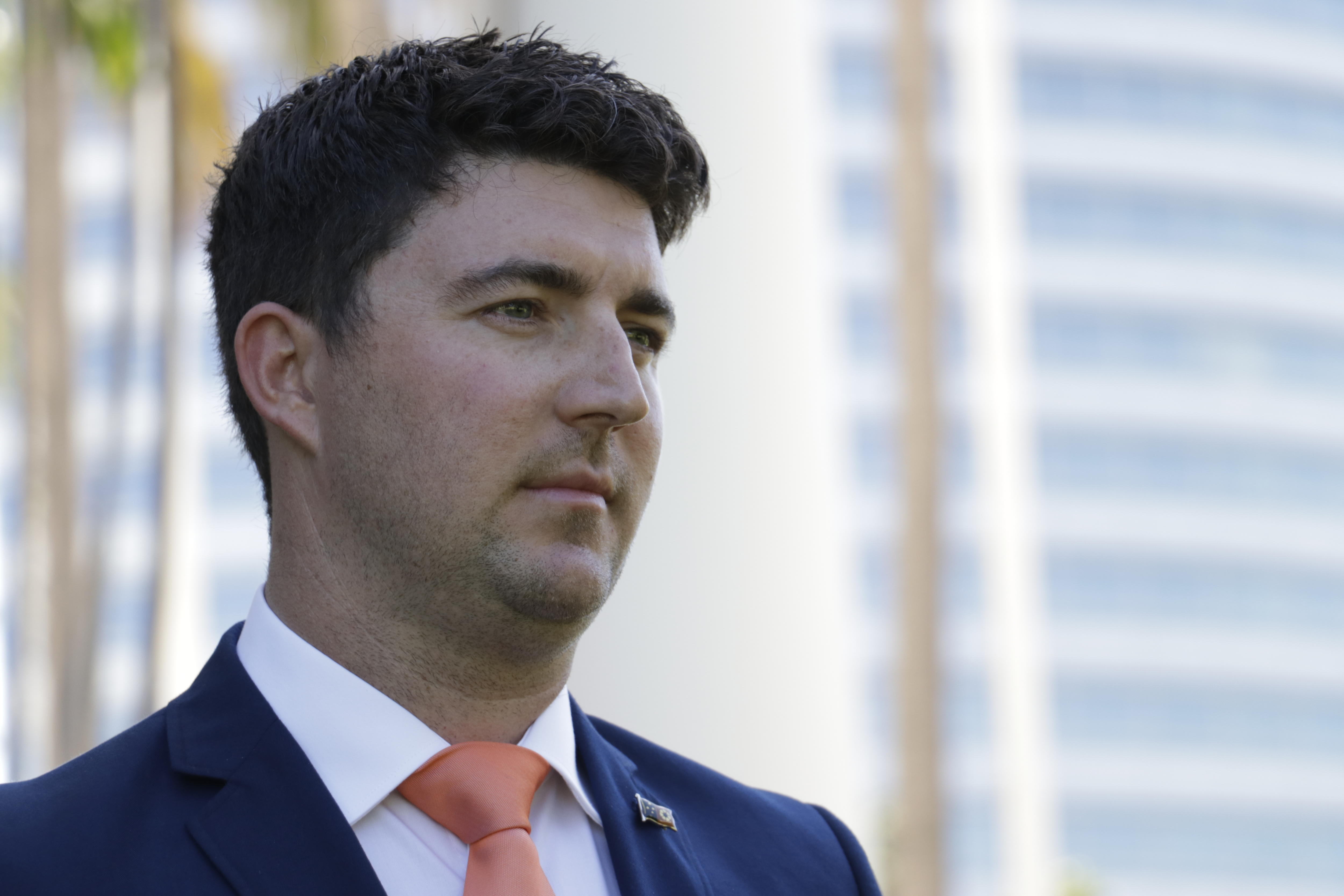 A close up photo of a man wearing a suit and tie with short brown hair. He looks serious, and is in his mid-30s.