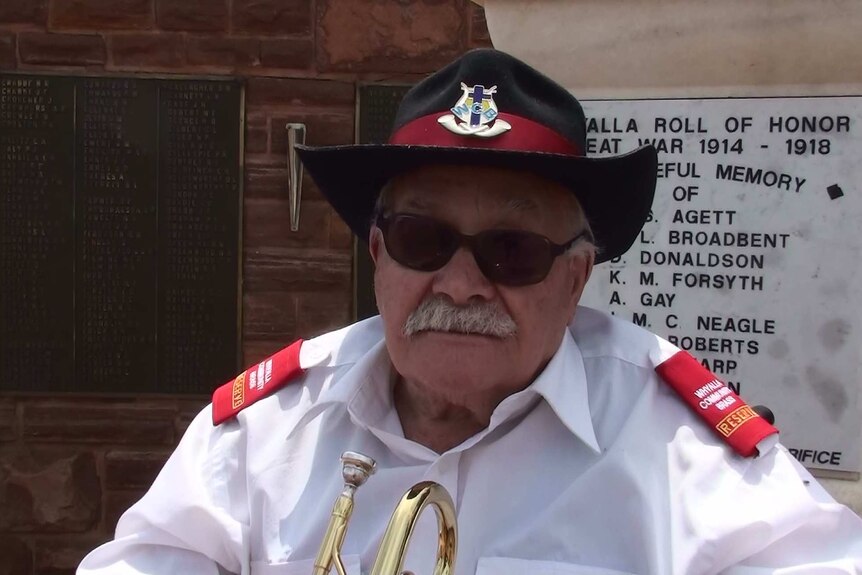 An old man wearing a black hat, white long-sleeve shirt and black sunglasses sits in front of the Whyalla Roll of Honour.