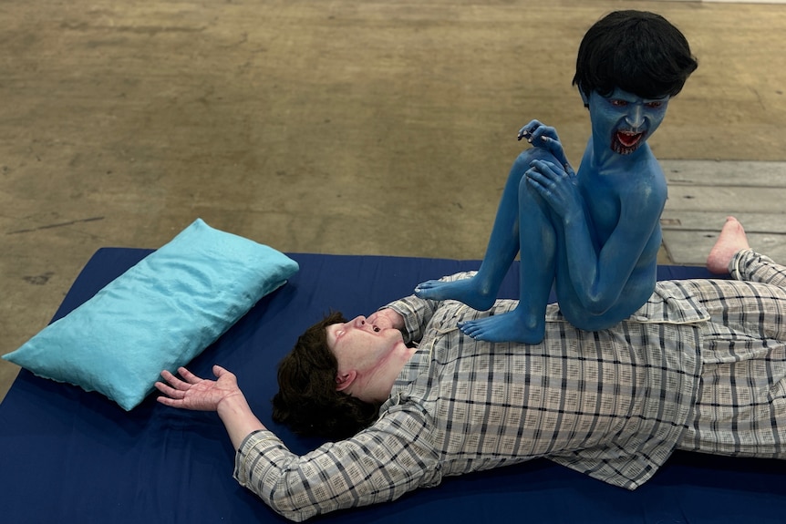 Una escultura de un hombre acostado sobre un colchón azul con una almohada cerca.  Una pequeña criatura azul parecida a un niño está sentada sobre su pecho.