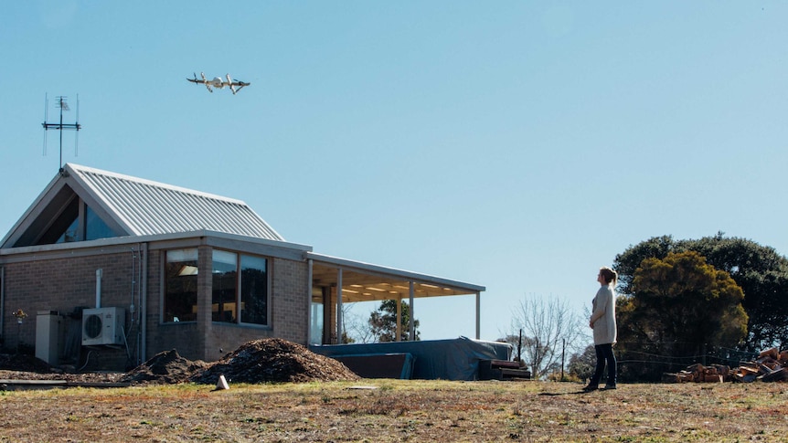 A woman stand in her yard as a drone hovers over her house.
