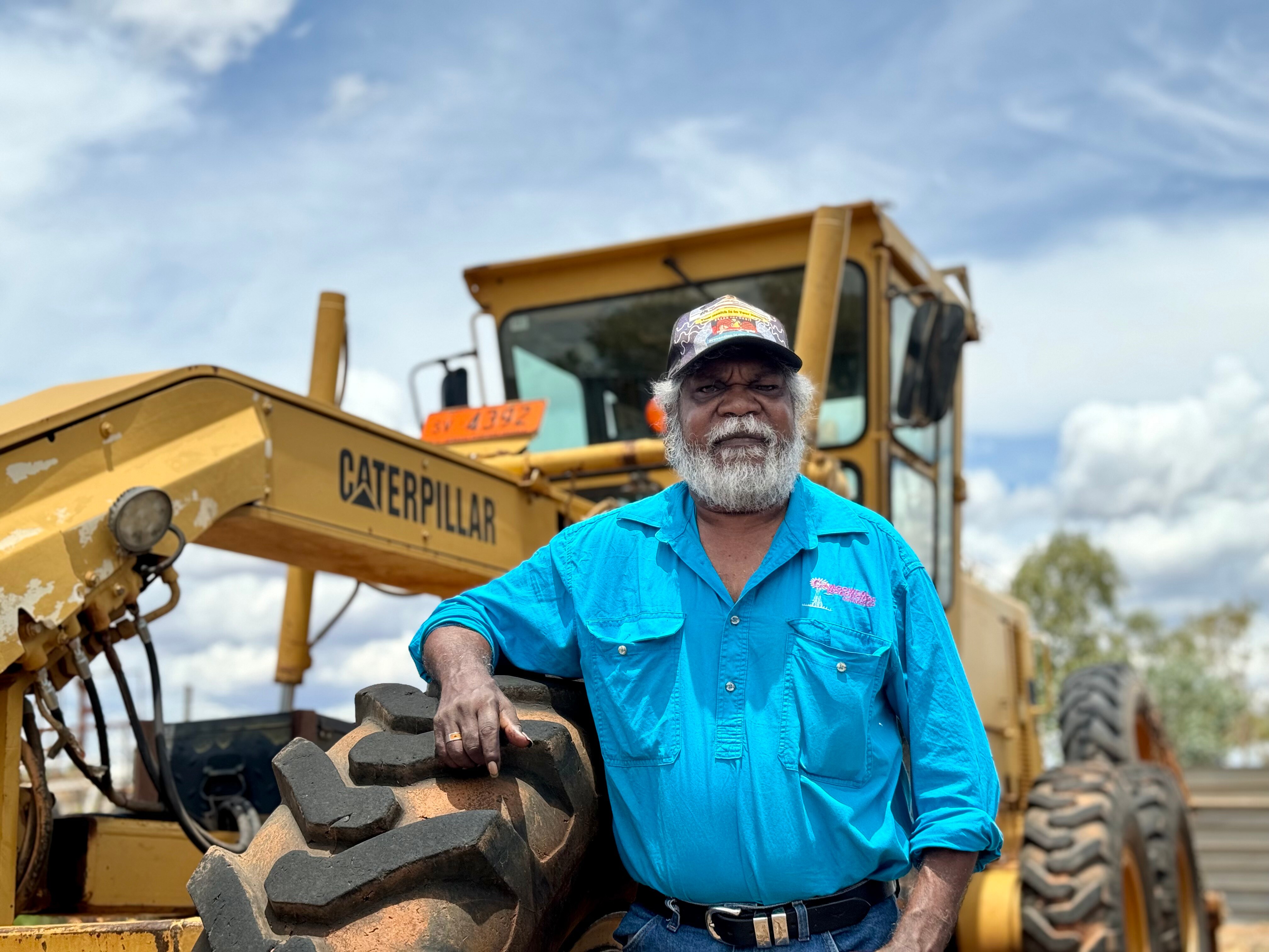An Aboriginal man in a light blue work shirt looks down slightly at the camera as he rests his arm on a grader front wheel