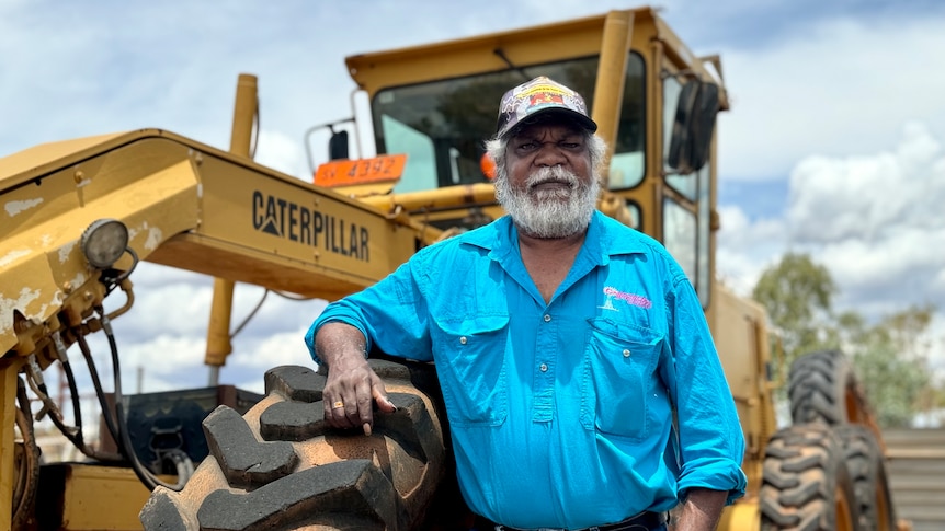 An Aboriginal man in a light blue work shirt looks down slightly at the camera as he rests his arm on a grader front wheel