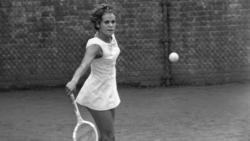 A black and white photo of a young woman staring at a tennis ball that she is about to hit.