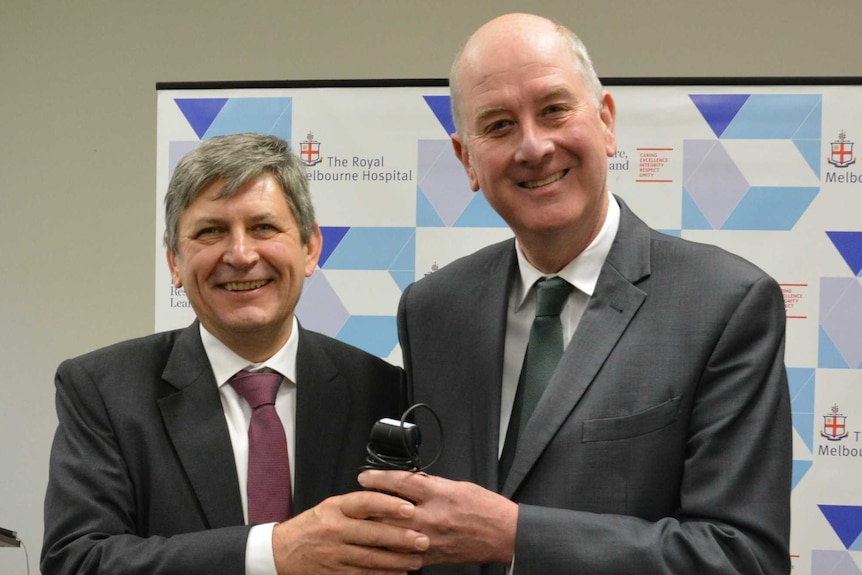 Surgeon Alistair Royse stands next to Richard Wynne, both in suits, as the hold a portable medical device and smile.