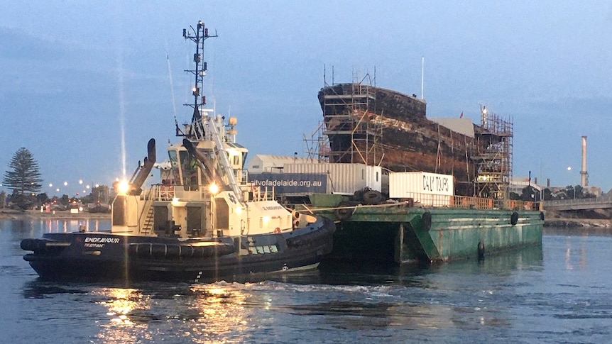 The hull of an aging ship sits on a barge and is pushed by a tug boat.