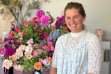 A woman in a white shirt smiles at the camera in front of a number of bunches of flowers