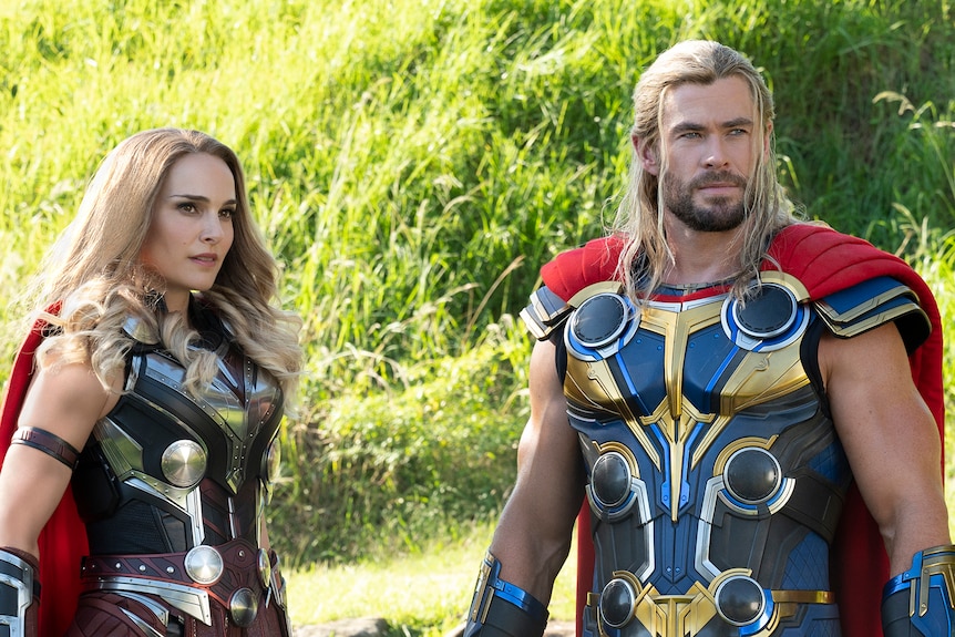White woman with curled blonde hair stands with man with long blonde hair. They both wear blue and gold armor and a red cape.