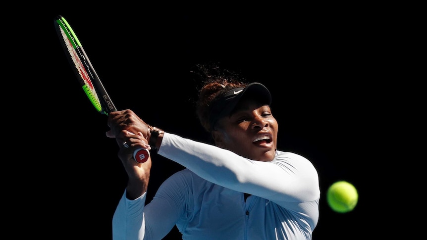 Serena Williams wears a white long-sleeve shirt and black visor as she leans in to hit tennis ball with racket in her hands.