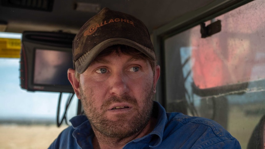 A man wearing a blue shirt, brown cap, with stubble and blue eyes, sits in the cabin of a header.