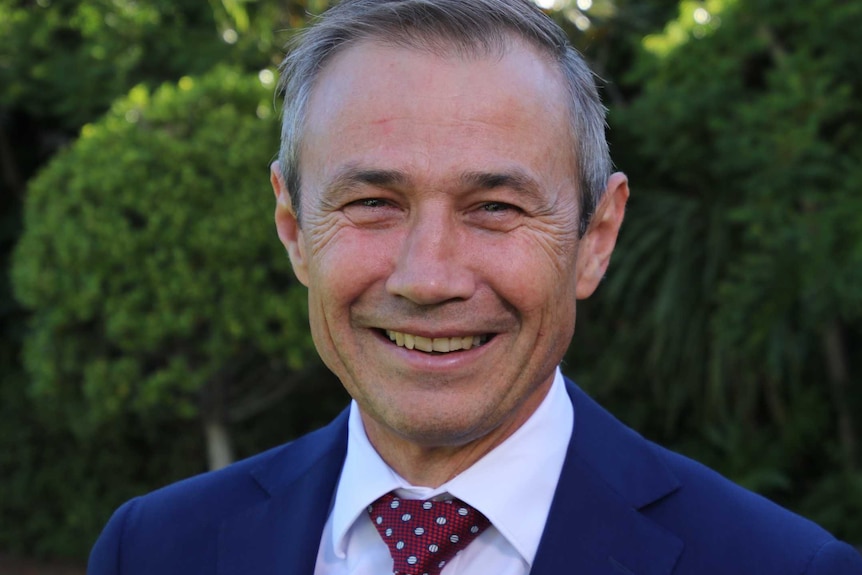 A headshot of Roger Cook smiling and wearing a blue suit and red tie standing outside.