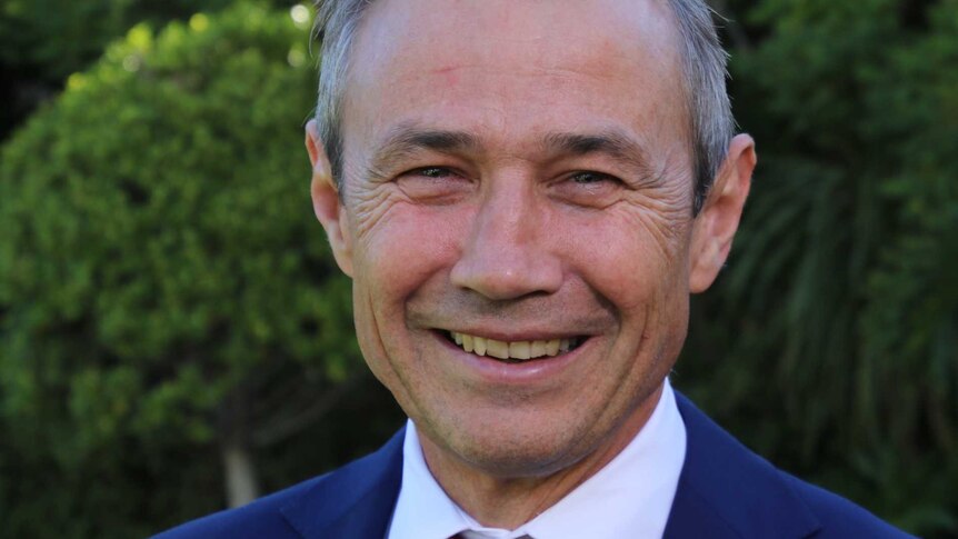 A headshot of Roger Cook smiling and wearing a blue suit and red tie standing outside.