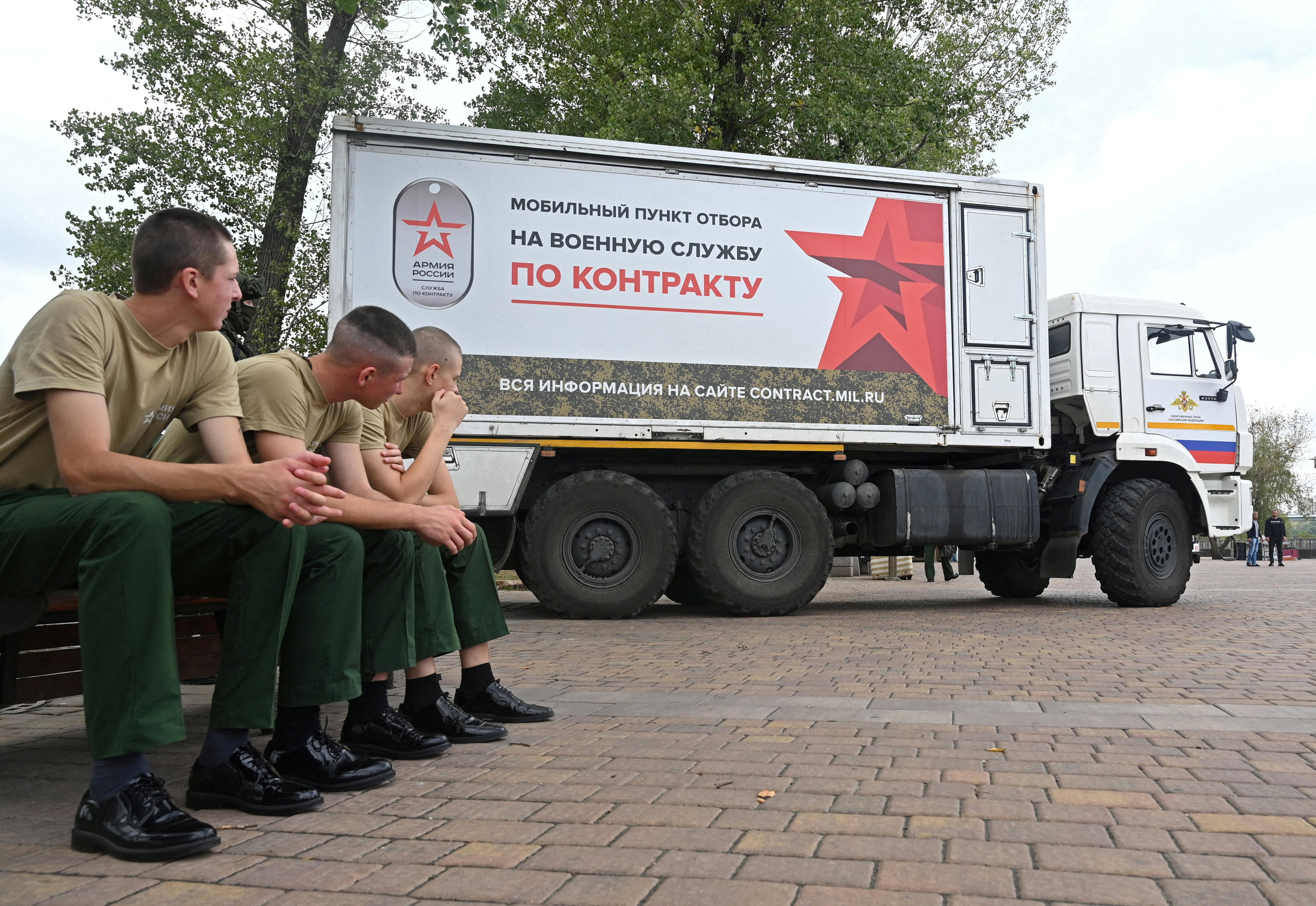 Three men in uniforms sit on a seat outside watching a truck that advertises in red writing and a red star.