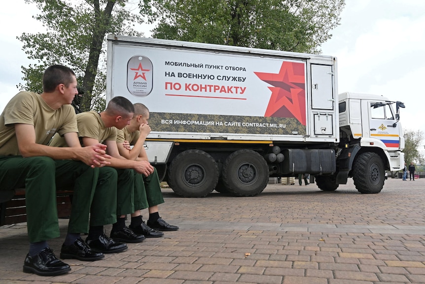 Three men in uniforms sit on a seat outside watching a truck that advertises in red writing and a red star.