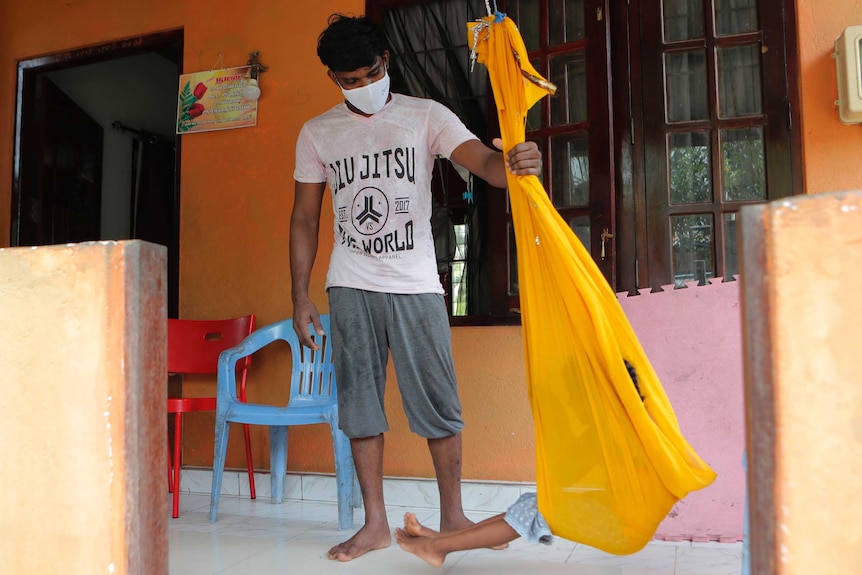 A man in shorts, t-shirt and face mask swings a small child on a hammock