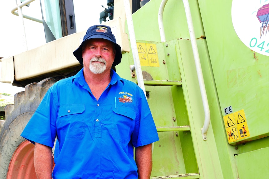 A bearded man in a hat standing in front of a harvester.