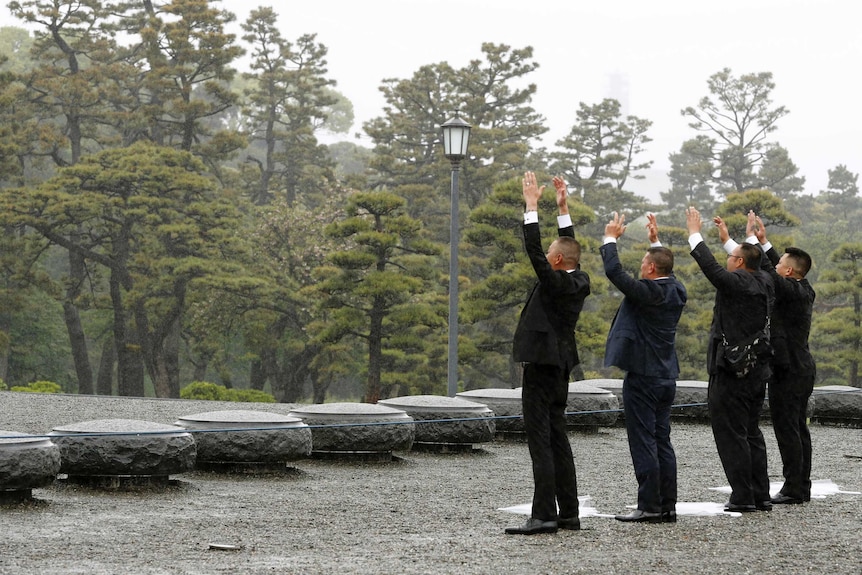 A group of four men in dark suits stand in a line and raise their hands in as a lush garden is behind them.