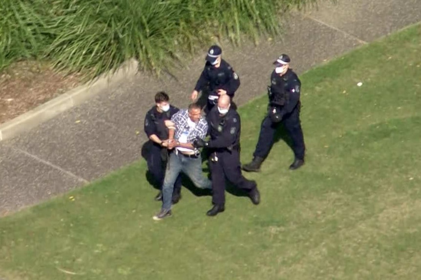 A still from aerial footage shows a man being arrested by police on a green lawn.
