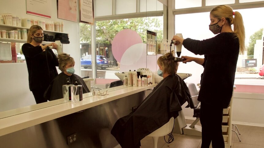 A woman dries another woman's hair in a salon with their images reflected in a mirror.