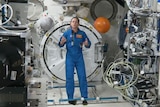 A female astronaut in a blue jumpsuit stands inside a space craft talking to an audience. 