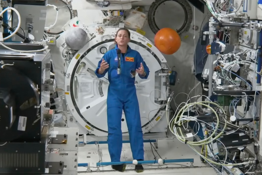A female astronaut in a blue jumpsuit stands inside a space craft talking to an audience. 