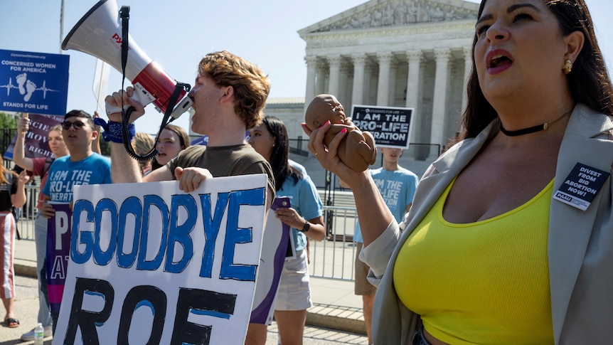 A group of protesters, one holding a sign that says GOODBYE ROE and shouting into a megaphone, outside the Supreme Court