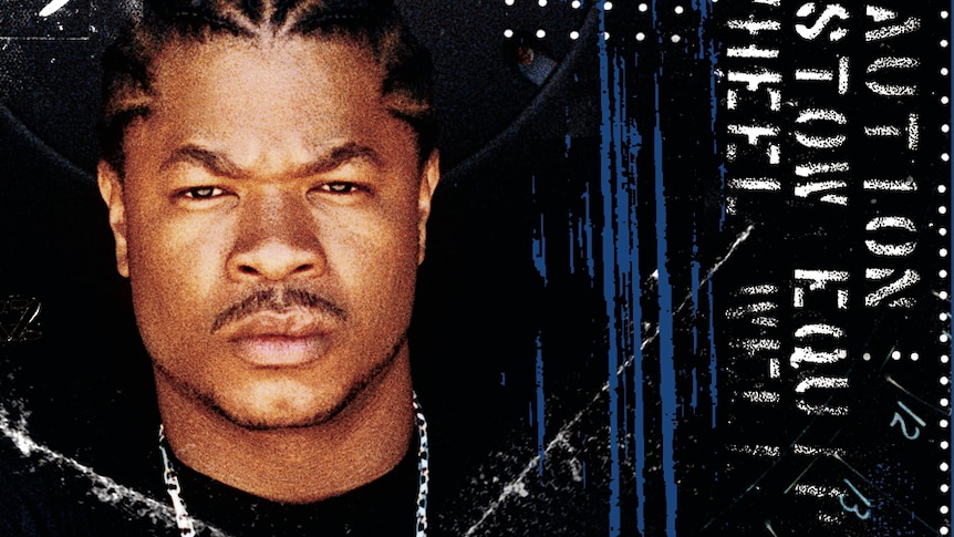 Rapper Xzibit holds his forearm up as a sign of strength. He has a stern look on his face. 