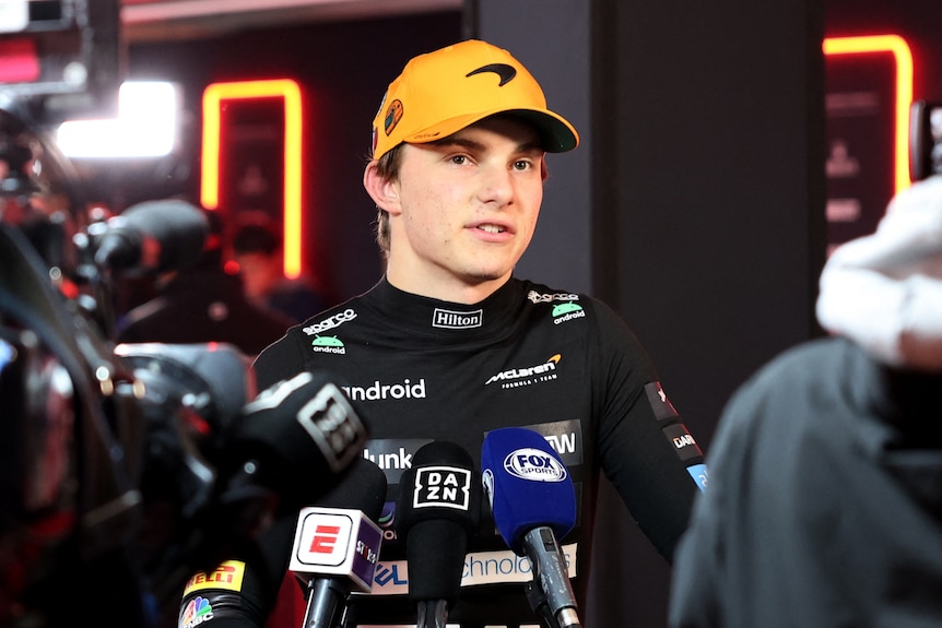 An F1 driver, in his race suit and a cap, speaking to media in front of microphones.