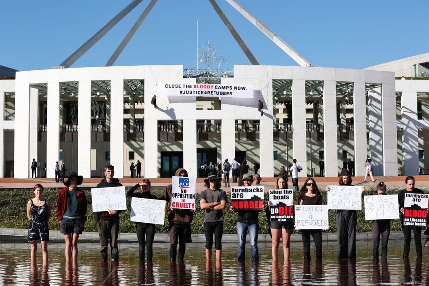 Protesters stand outside Parliament House with signs as two men abseil down the front of the building to unfurl a banner