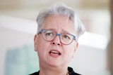 A woman with short grey hair and glasses talking to media