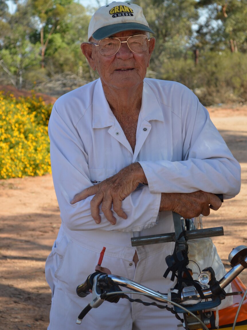 An older man wearing white overalls on a bicycle with scrub in the background.