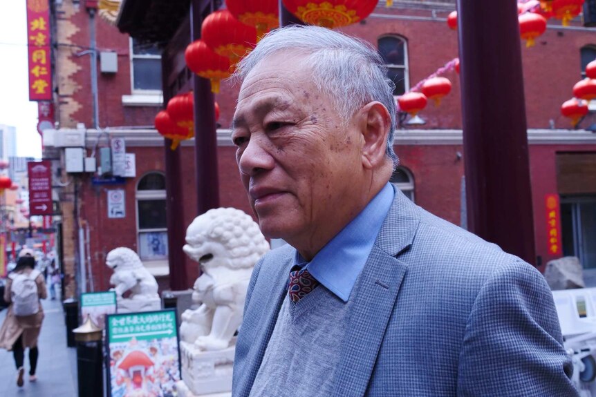 An elderly Chinese man stands in the main square of Melbourne's China town in front of red lanterns and white stone dragon
